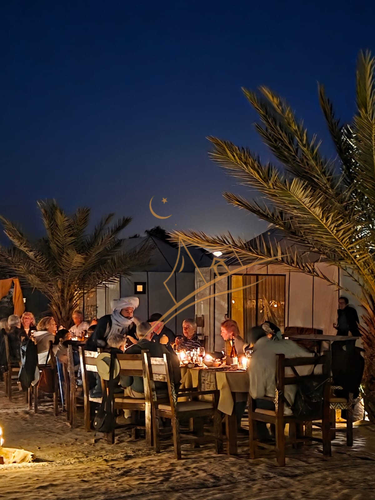 Guests dining at a long candlelit table beneath the palms at dusk