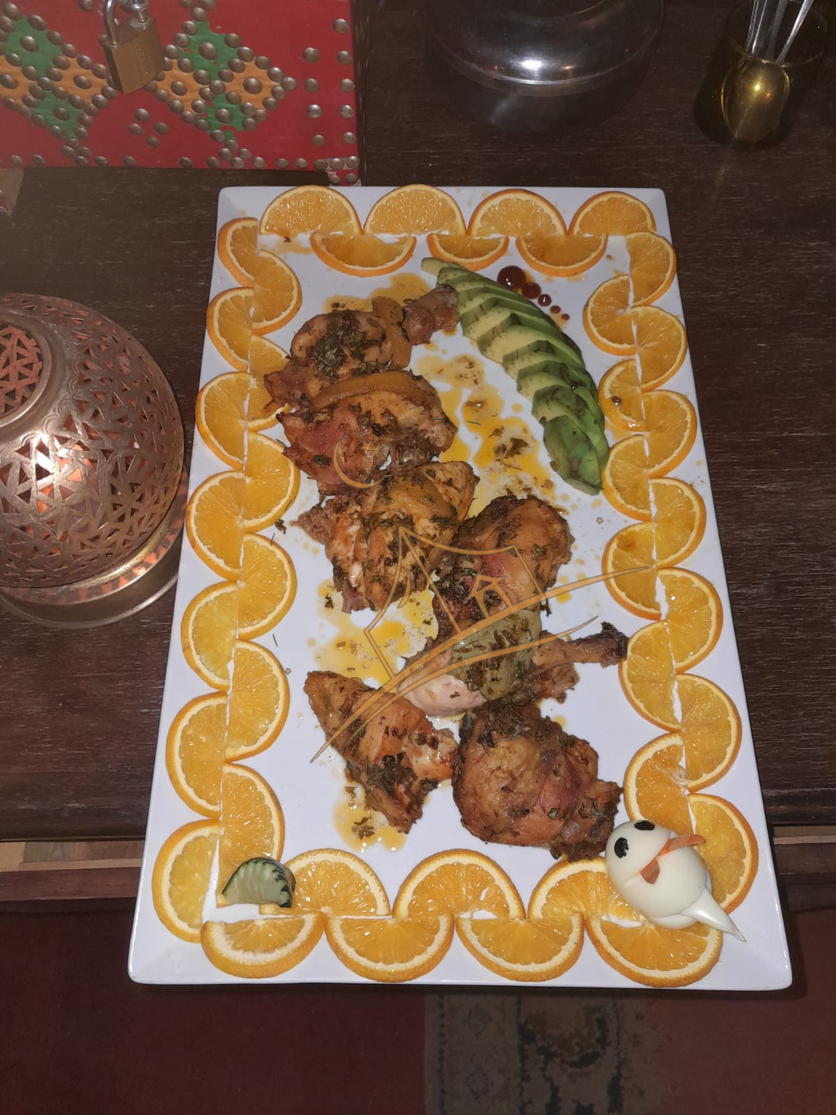 Herb-roasted chicken plated with a fanned avocado and an orange-slice border beside a Moroccan lantern