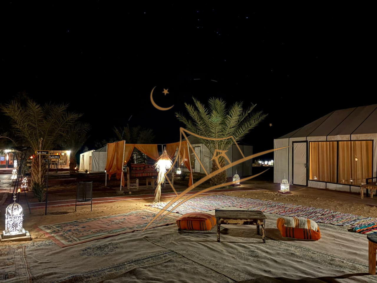 Outdoor lounge with cushions and rugs beneath a star-filled sky beside a glowing palm-frond lamp